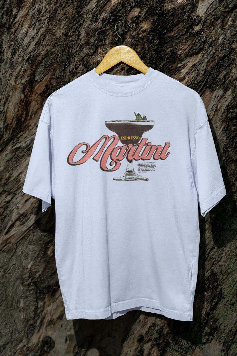 Puede incluir: Camiseta azul claro con un gr&aacute;fico de un espresso martini. El dise&ntilde;o incluye la palabra "Espresso" encima de la copa de martini y la palabra "Martini" en cursiva. La camiseta est&aacute; colgada en una percha de madera sobre un fondo texturizado.