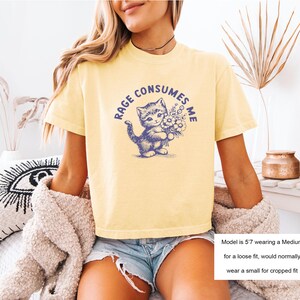 May include: Yellow t-shirt with a blue graphic of a kitten holding flowers and the text "RAGE CONSUMES ME". The shirt is a light yellow color.