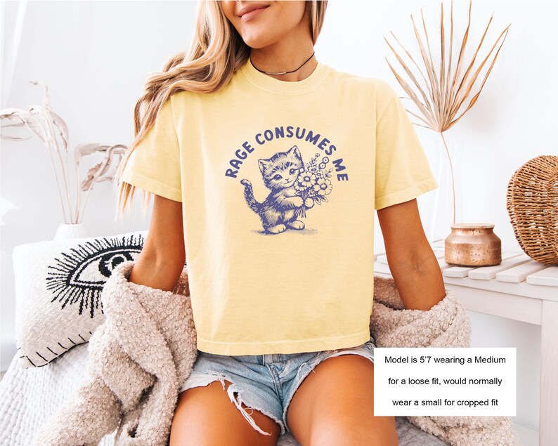 May include: Yellow t-shirt with a blue graphic of a kitten holding flowers and the text "RAGE CONSUMES ME". The shirt is a light yellow color.