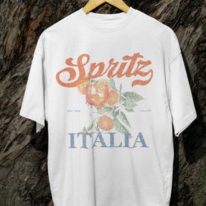 May include: White t-shirt with a vintage-style design. The design features the word "Spritz" in orange script above an illustration of oranges and leaves. Below the illustration, the word "Italia" is printed in blue.