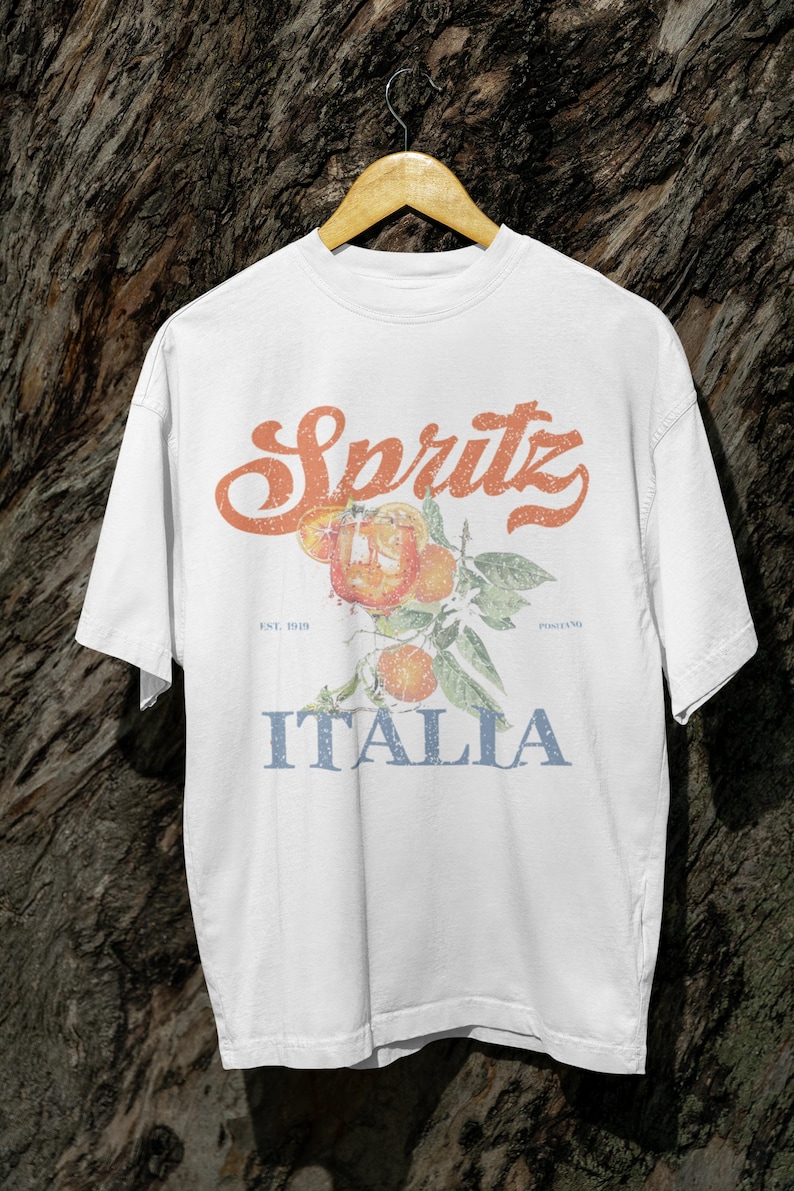 May include: White t-shirt with a vintage-style design. The design features the word "Spritz" in orange script above an illustration of oranges and leaves. Below the illustration, the word "Italia" is printed in blue.