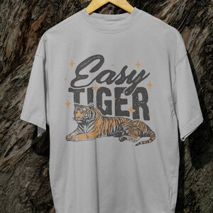 K&ouml;nnte beinhalten: Hellgraues T-Shirt mit einer Tiger-Illustration. Der Tiger ist orange und schwarz, liegt. Der Text "Easy Tiger" steht in einer verblassten Schriftart mit Sternen &uuml;ber dem Tiger. Das T-Shirt h&auml;ngt an einem Holzb&uuml;gel.
