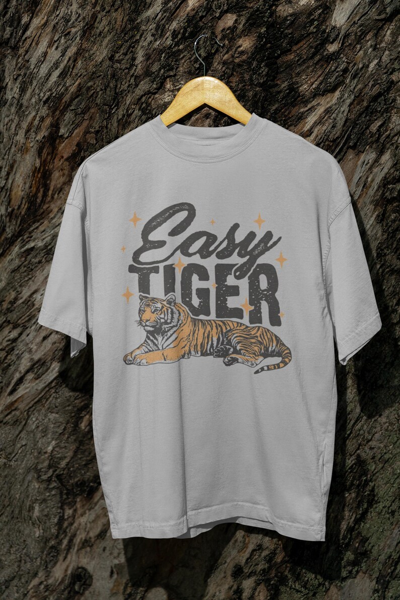 K&ouml;nnte beinhalten: Hellgraues T-Shirt mit einer Tiger-Illustration. Der Tiger ist orange und schwarz, liegt. Der Text "Easy Tiger" steht in einer verblassten Schriftart mit Sternen &uuml;ber dem Tiger. Das T-Shirt h&auml;ngt an einem Holzb&uuml;gel.
