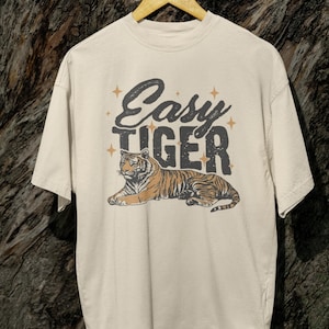 K&ouml;nnte beinhalten: Cremefarbenes T-Shirt mit einer Tiger-Illustration im Vintage-Stil. Der Text "Easy Tiger" ist in einer verwaschenen Schriftart mit Sternakzenten &uuml;ber dem Tiger aufgedruckt. Der Tiger ist in Orange- und Braunt&ouml;nen dargestellt.