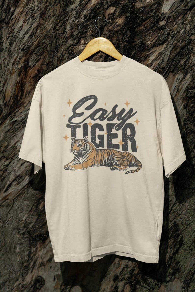 K&ouml;nnte beinhalten: Cremefarbenes T-Shirt mit einer Tiger-Illustration im Vintage-Stil. Der Text "Easy Tiger" ist in einer verwaschenen Schriftart mit Sternakzenten &uuml;ber dem Tiger aufgedruckt. Der Tiger ist in Orange- und Braunt&ouml;nen dargestellt.