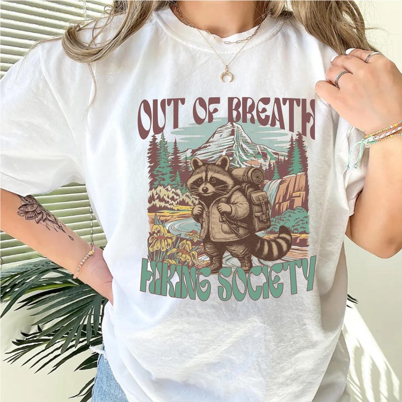 May include: White t-shirt with a graphic featuring a raccoon with a backpack, standing in front of a mountain and waterfall scene. Text reads "OUT OF BREATH" above and "HIKING SOCIETY" below the raccoon illustration.