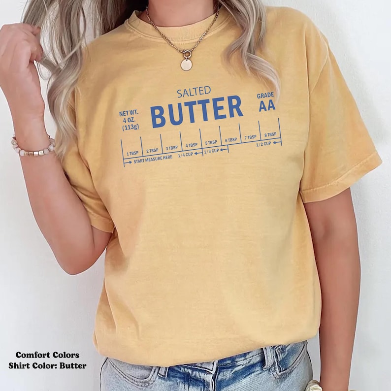 May include: Mustard yellow t-shirt with the words "SALTED BUTTER" in blue, along with a measurement guide. The shirt also has the text "GRADE AA" and "NET WT. 4 OZ. (113g)".