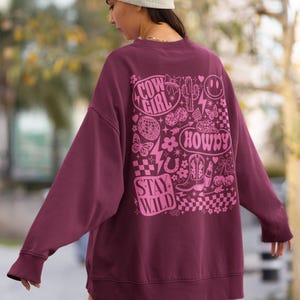 May include: A maroon sweatshirt with a pink graphic design on the back. The design includes the words "Cow Girl", "Howdy", and "Stay Wild", along with images of a cactus, boots, and a horseshoe.