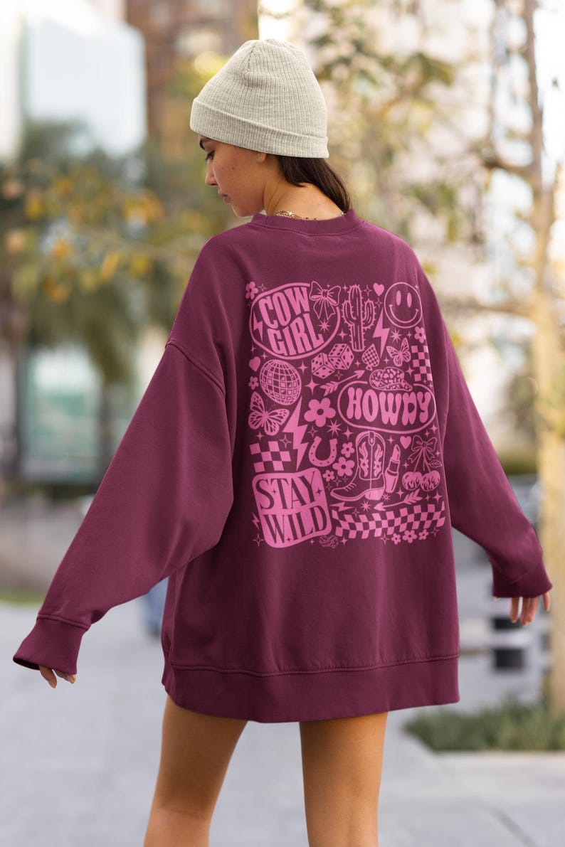 May include: A maroon sweatshirt with a pink graphic design on the back. The design includes the words "Cow Girl", "Howdy", and "Stay Wild", along with images of a cactus, boots, and a horseshoe.