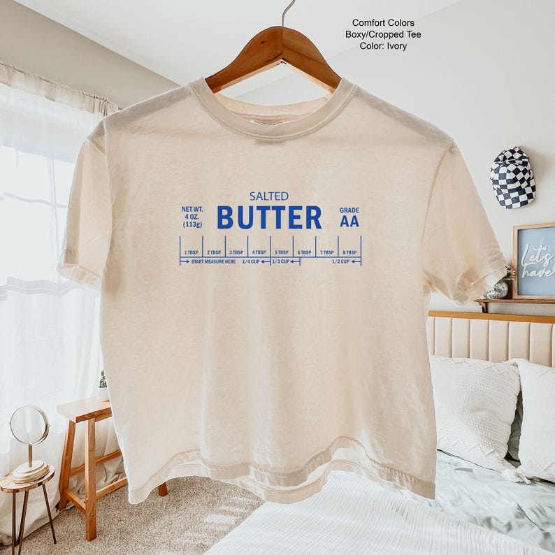 Puede incluir: Camiseta corta y cuadrada de color marfil con las palabras "SALTED BUTTER" en azul, junto con una gu&iacute;a de medici&oacute;n de mantequilla. La camiseta tambi&eacute;n tiene el texto "NET WT. 4 OZ. (113g)" y "GRADE AA".