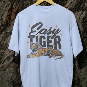 K&ouml;nnte beinhalten: Hellblaues T-Shirt mit dem Text "Easy Tiger" &uuml;ber einer Tigergrafik. Der Tiger ist orange und schwarz. Das T-Shirt h&auml;ngt an einem Holzb&uuml;gel.