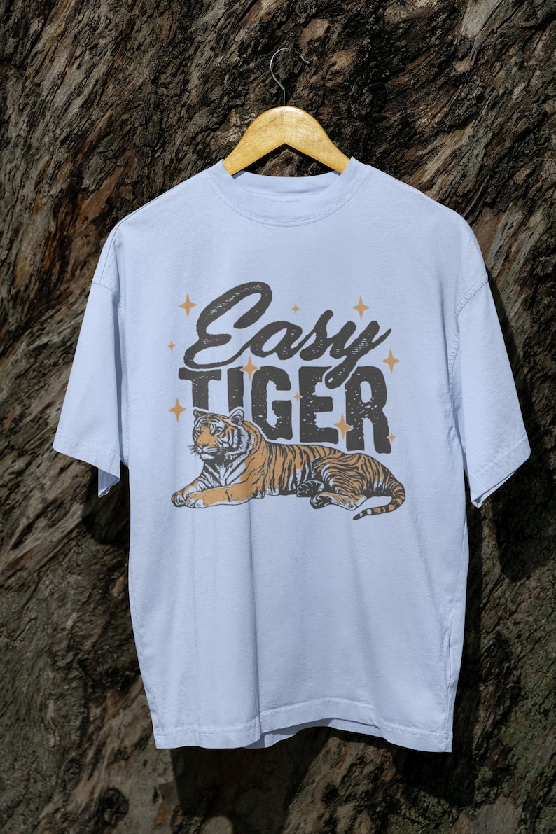 K&ouml;nnte beinhalten: Hellblaues T-Shirt mit dem Text "Easy Tiger" &uuml;ber einer Tigergrafik. Der Tiger ist orange und schwarz. Das T-Shirt h&auml;ngt an einem Holzb&uuml;gel.