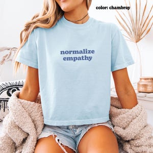 May include: Light blue t-shirt with the words "normalize empathy" in navy blue. The shirt is short-sleeved and made of a soft material. The color is chambray.