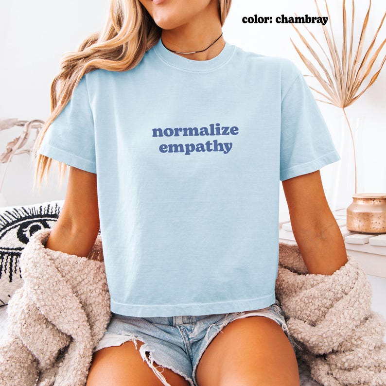 May include: Light blue t-shirt with the words "normalize empathy" in navy blue. The shirt is short-sleeved and made of a soft material. The color is chambray.