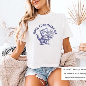 May include: White t-shirt with a blue illustration of a kitten holding flowers, and the text "RAGE CONSUMES ME" in a circular design. The shirt is a casual style.