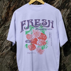 May include: A lavender t-shirt with a graphic print of raspberries and the text "FRESH Raspberries" in a vintage style font. The text "ENJOY LITTLE THINGS" is printed on the right side of the graphic. The text "SWEET AND FRESH" is printed below the graphic.