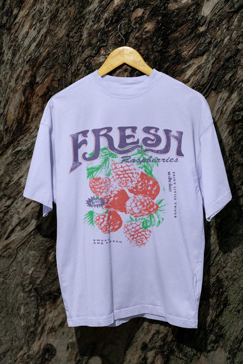 May include: A lavender t-shirt with a graphic print of raspberries and the text "FRESH Raspberries" in a vintage style font. The text "ENJOY LITTLE THINGS" is printed on the right side of the graphic. The text "SWEET AND FRESH" is printed below the graphic.
