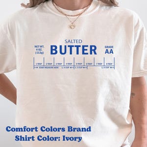May include: Ivory-colored t-shirt with a blue graphic resembling a butter package, including measurements in tablespoons and cups. The text reads "SALTED BUTTER" and "GRADE AA". The shirt also displays "Comfort Colors Brand" and "Shirt Color: Ivory".