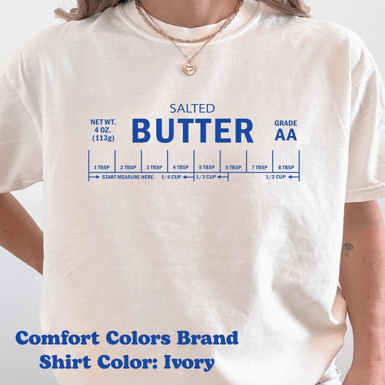 May include: Ivory-colored t-shirt with a blue graphic resembling a butter package, including measurements in tablespoons and cups. The text reads "SALTED BUTTER" and "GRADE AA". The shirt also displays "Comfort Colors Brand" and "Shirt Color: Ivory".