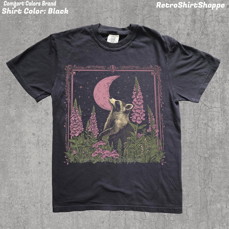 May include: Black t-shirt with a raccoon graphic looking up at a pink crescent moon. The design includes pink flowers and the text "RetroShirtShoppe". The shirt is from the Comfort Colors Brand.