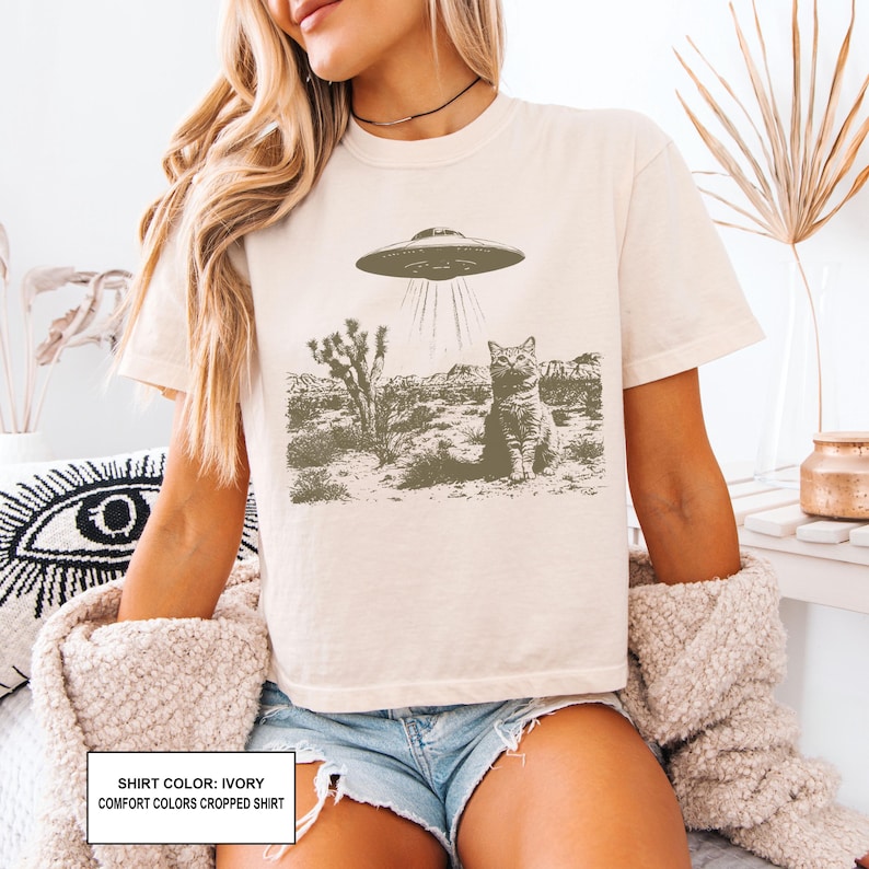 May include: Ivory-colored t-shirt featuring a graphic of a cat being beamed up by a UFO in a desert landscape. The shirt has short sleeves and a crew neck. The text on the shirt reads "SHIRT COLOR: IVORY COMFORT COLORS CROPPED SHIRT".