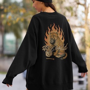 May include: Black sweatshirt with a graphic of a cheetah holding an 8-ball, surrounded by flames. The text "Stay Wild" is below the cheetah. A pair of dice and a matchbox are also in the design. The sweatshirt has long sleeves and a crew neck.
