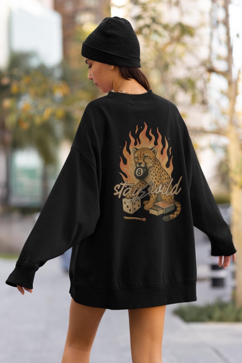 May include: Black sweatshirt with a graphic of a cheetah holding an 8-ball, surrounded by flames. The text "Stay Wild" is below the cheetah. A pair of dice and a matchbox are also in the design. The sweatshirt has long sleeves and a crew neck.