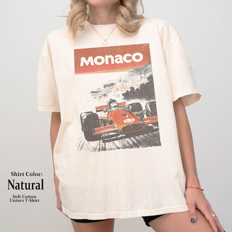以下が含まれることがあります： ナチュラルカラーのユニセックスTシャツ。レトロなレーシングカーのグラフィックと、赤いボックスに「モナコ」の文字が入っています。柔らかい綿で作られたシャツです。