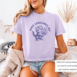 May include: A light purple t-shirt with the text "RAGE CONSUMES ME" surrounding a blue illustration of a kitten holding flowers. The shirt is short-sleeved and has a relaxed fit.