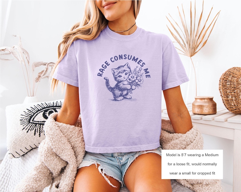 May include: A light purple t-shirt with the text "RAGE CONSUMES ME" surrounding a blue illustration of a kitten holding flowers. The shirt is short-sleeved and has a relaxed fit.