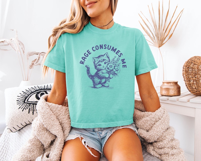 May include: A turquoise t-shirt with a round graphic featuring a kitten holding flowers and the text "RAGE CONSUMES ME" in a circle. The shirt is made of a soft material.