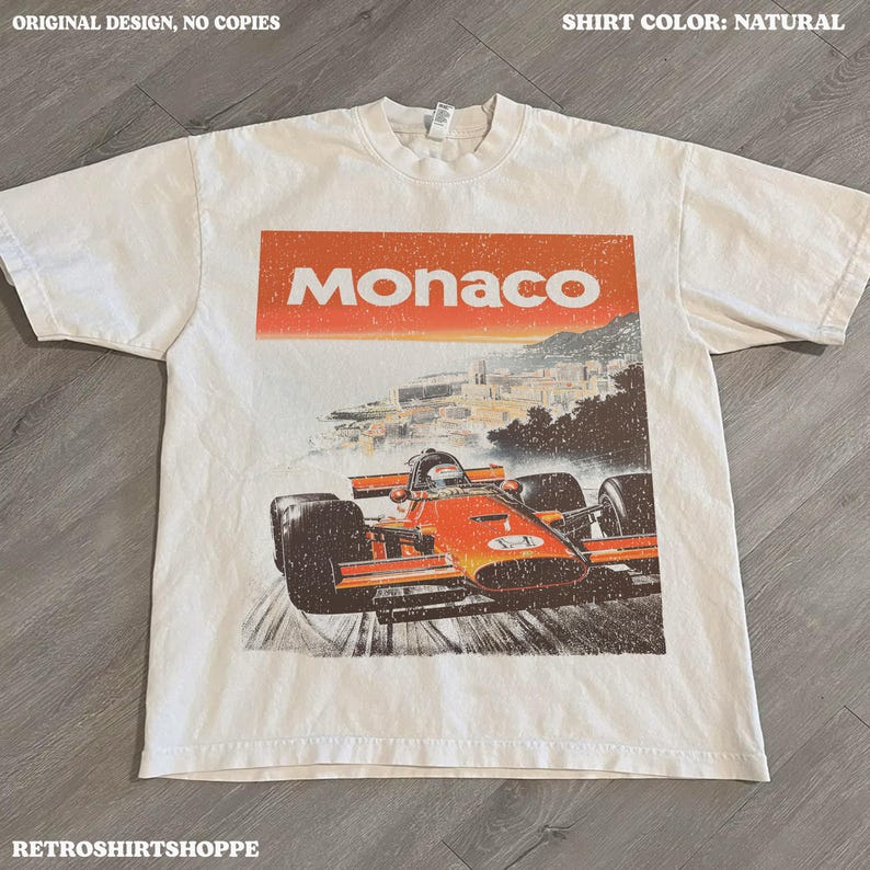 以下が含まれることがあります： ナチュラルカラーのTシャツ。オレンジ色の長方形に「Monaco」の文字と、F1レーシングカーのレトロなグラフィックがプリントされています。デザインには、海岸沿いの都市の背景が含まれています。「ORIGINAL DESIGN, NO COPIES」の文字が上部にあります。