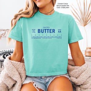 Puede incluir: Una camiseta verde menta con la palabra "BUTTER" en azul, junto con una gu&iacute;a de medici&oacute;n de mantequilla. La camiseta tambi&eacute;n presenta el texto "SALTED", "NET WT. 113g" y "GRADE AA".