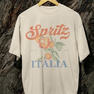 Op de afbeelding: Crèmekleurig T-shirt met een vintage-stijl grafische afbeelding. Het ontwerp toont het woord "Spritz" in oranje script boven een illustratie van sinaasappels en bladeren. Daaronder staat het woord "Italia" in blauw gedrukt.