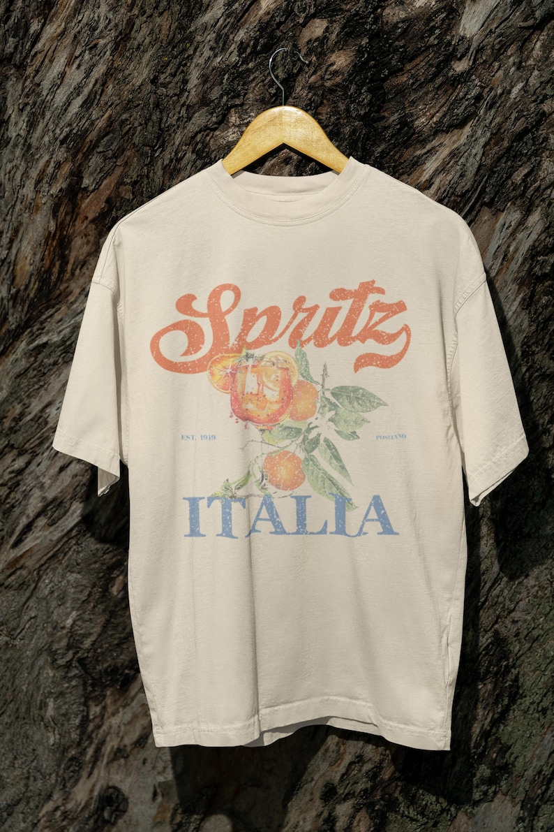May include: Cream-colored t-shirt with a vintage-style graphic. The design features the word "Spritz" in orange script above an illustration of oranges and leaves. Below the illustration, the word "Italia" is printed in blue.