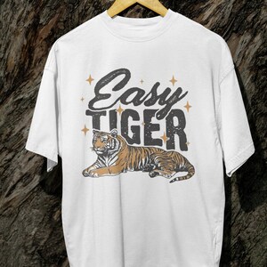 K&ouml;nnte beinhalten: Wei&szlig;es T-Shirt mit dem Text "Easy Tiger" im Vintage-Stil, mit einer Tiger-Illustration. Der Tiger ist braun und schwarz und liegt. Das Design enth&auml;lt Sterne.