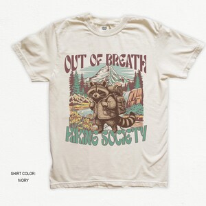 May include: Ivory-colored t-shirt featuring a raccoon with a backpack in a mountain scene. The text "OUT OF BREATH" is above the raccoon, and "HIKING SOCIETY" is below. The shirt is a crew neck.
