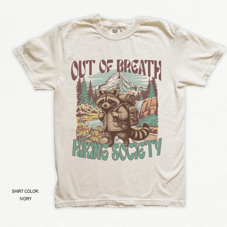 May include: Ivory-colored t-shirt featuring a raccoon with a backpack in a mountain scene. The text "OUT OF BREATH" is above the raccoon, and "HIKING SOCIETY" is below. The shirt is a crew neck.
