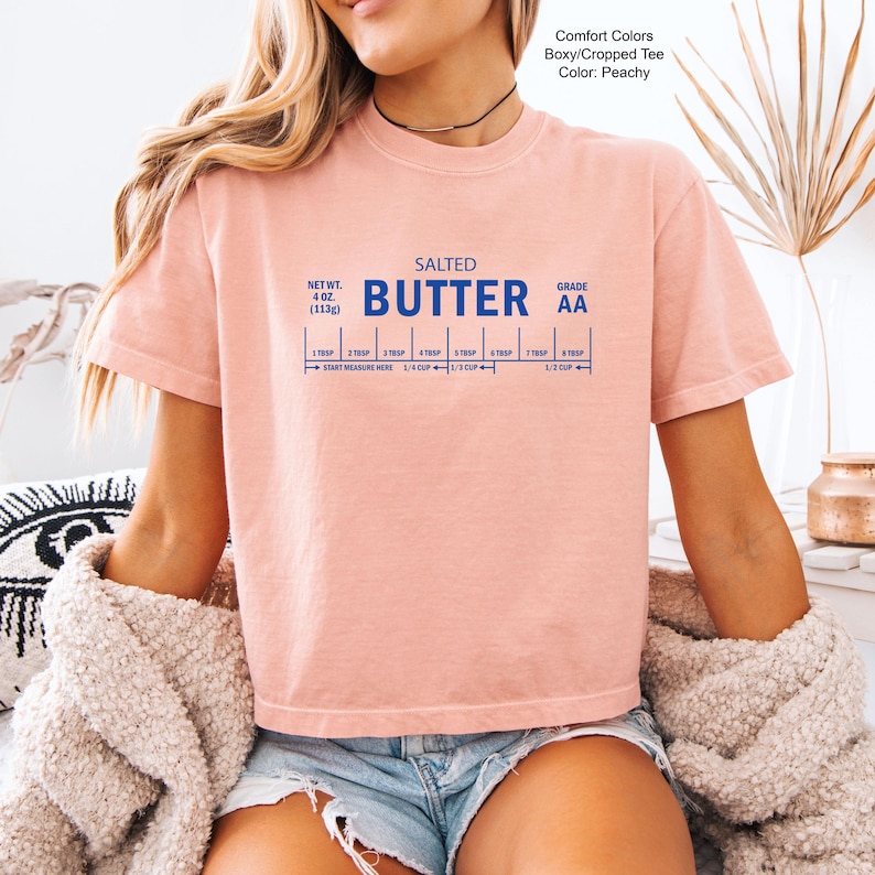 Puede incluir: Camiseta corta y holgada de color melocot&oacute;n con las palabras "SALTED BUTTER" en azul. Debajo, una gu&iacute;a de medidas azul. La camiseta tambi&eacute;n tiene el texto "NET WT. 4 OZ (113g)" y "GRADE AA".