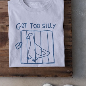 May include: Pale blue t-shirt featuring a blue drawing of a bird in a cage and the words "GOT TOO SILLY". The shirt is folded on a wooden surface, with white trainers in the foreground.