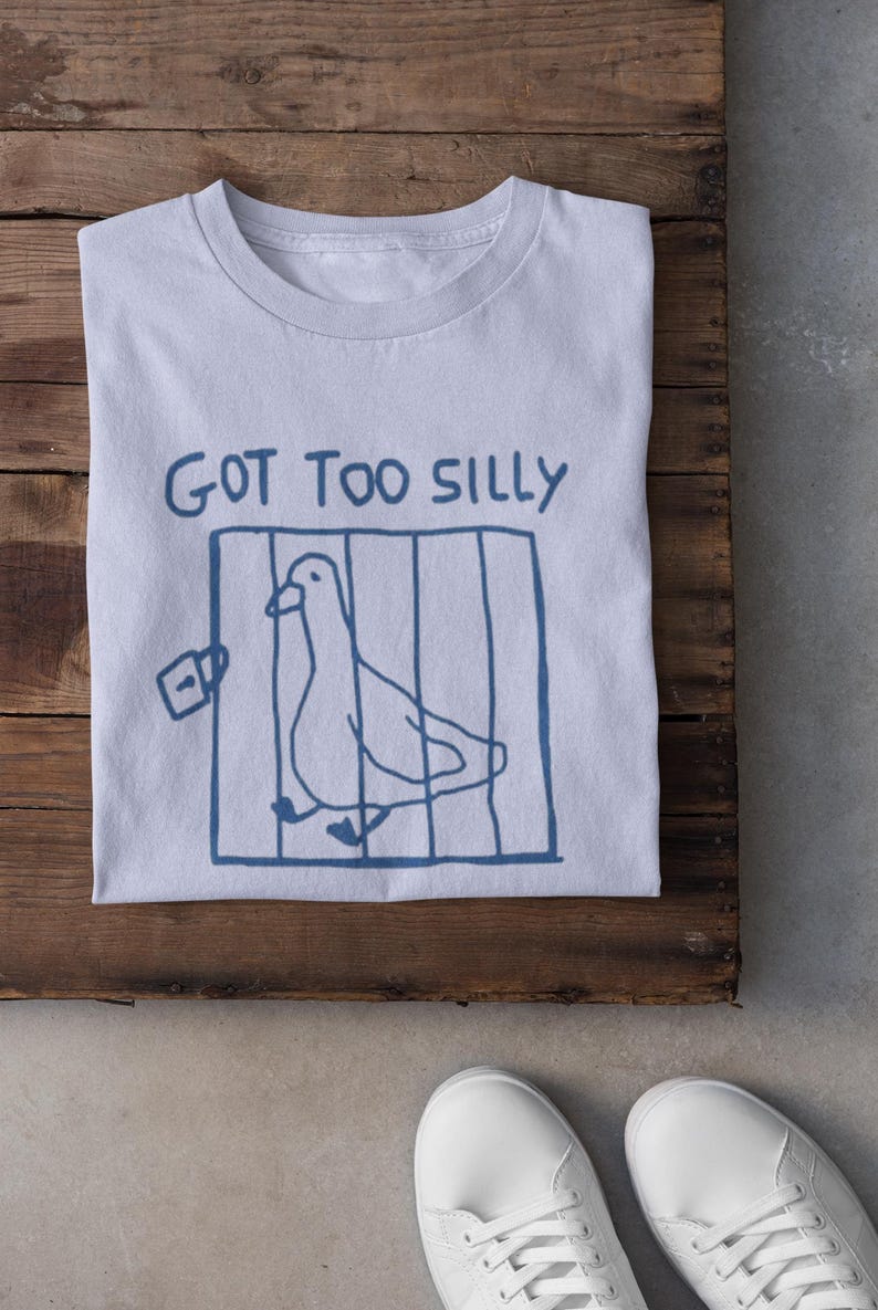 May include: Pale blue t-shirt featuring a blue drawing of a bird in a cage and the words "GOT TOO SILLY". The shirt is folded on a wooden surface, with white trainers in the foreground.