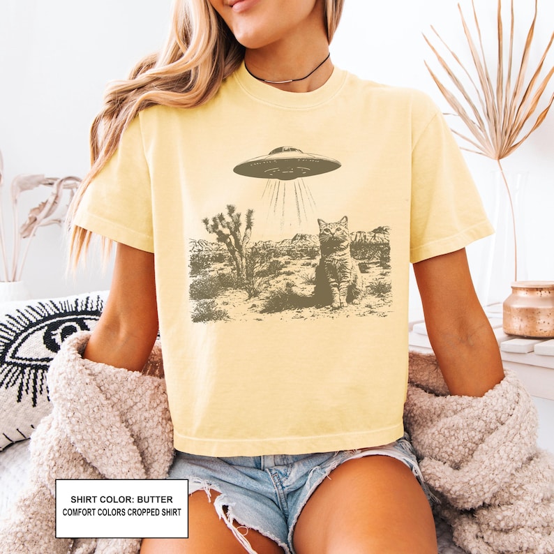 May include: Butter-colored t-shirt featuring a desert landscape with a cat and a UFO. The graphic is in shades of brown and gray. The shirt has short sleeves and a crew neck. The text on the shirt reads "SHIRT COLOR: BUTTER COMFORT COLORS CROPPED SHIRT".