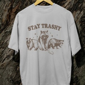 K&ouml;nnte beinhalten: Ein wei&szlig;es T-Shirt mit einer braunen Grafik von drei Waschb&auml;ren, die Sonnenbrillen tragen. Der Text "STAY TRASHY" ist in Braun &uuml;ber den Waschb&auml;ren gedruckt.