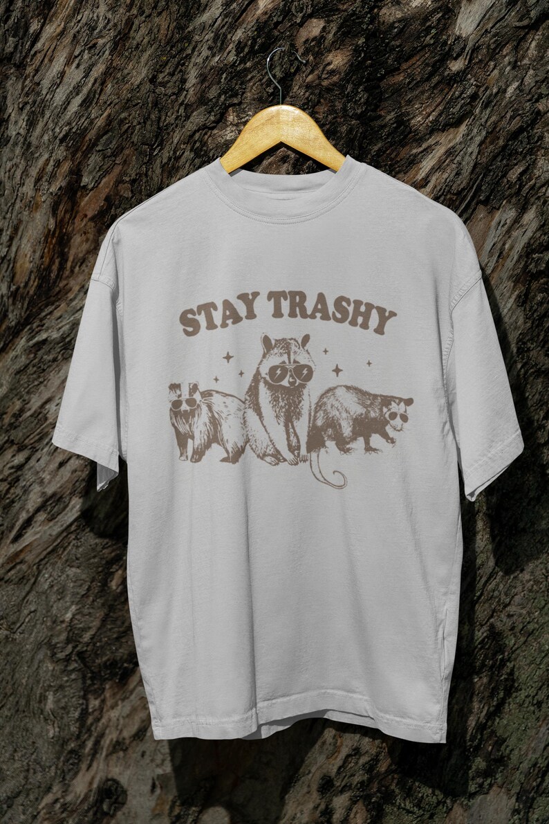K&ouml;nnte beinhalten: Ein wei&szlig;es T-Shirt mit einer braunen Grafik von drei Waschb&auml;ren, die Sonnenbrillen tragen. Der Text "STAY TRASHY" ist in Braun &uuml;ber den Waschb&auml;ren gedruckt.