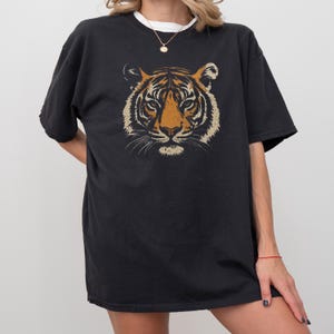 K&ouml;nnte beinhalten: Schwarzes T-Shirt mit einem Tigergesicht-Motiv. Das Tigergesicht ist mit orange, beige und wei&szlig;en Farben detailliert. Das T-Shirt hat kurze &Auml;rmel und eine lockere Passform, geeignet f&uuml;r den t&auml;glichen Gebrauch.