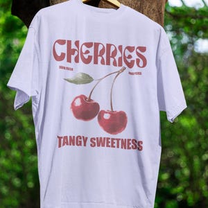 May include: White t-shirt with a cherry graphic. The word "CHERRIES" is printed in a retro font above a pair of red cherries with a green leaf. Below the cherries is the phrase "TANGY SWEETNESS".