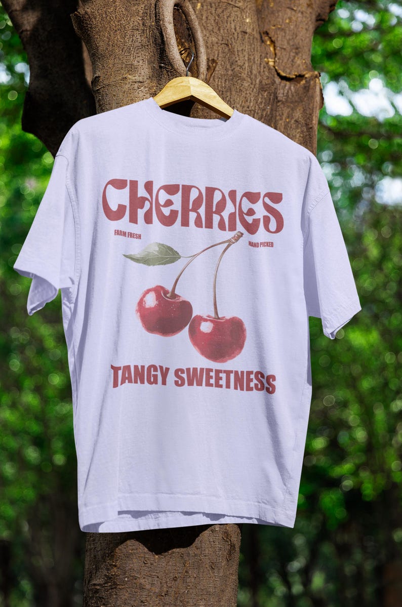 May include: White t-shirt with a cherry graphic. The word "CHERRIES" is printed in a retro font above a pair of red cherries with a green leaf. Below the cherries is the phrase "TANGY SWEETNESS".