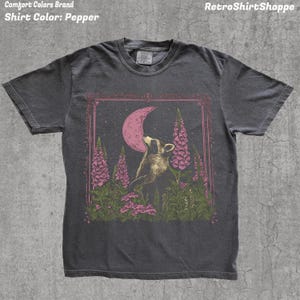 May include: Pepper-colored t-shirt featuring a raccoon gazing at a pink crescent moon within a decorative frame. The design includes pink flowers and foliage. The text "RetroShirtShoppe" is at the top right.