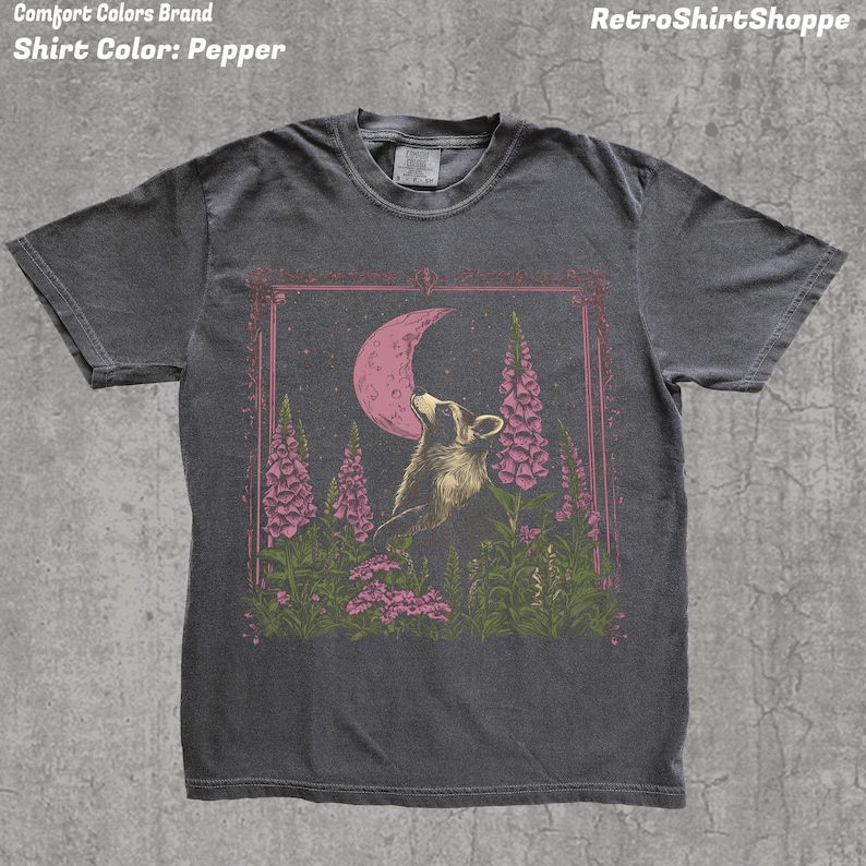 May include: Pepper-colored t-shirt featuring a raccoon gazing at a pink crescent moon within a decorative frame. The design includes pink flowers and foliage. The text "RetroShirtShoppe" is at the top right.