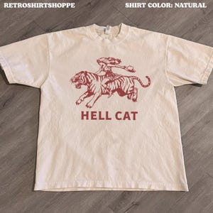 Könnte beinhalten: Naturfarbenes T-Shirt mit einem weinroten Aufdruck einer Frau, die auf einem Tiger reitet, und dem Text "HELL CAT" darunter. Das Shirt hat einen Rundhalsausschnitt und kurze Ärmel. Der Markenname "RETROSHIRTSHOPPE" steht oben links.